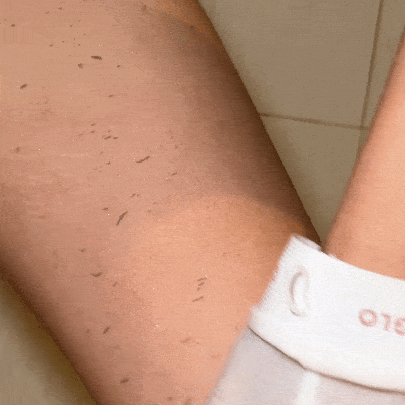 Close-up image of someone's lower leg during a shaving process using the CheekyGlo Sensitive Exfoliating Glove. Small hair clippings are visible on the leg and floor. The person is wearing white socks with a red logo and is sitting on what appears to be a tiled floor.